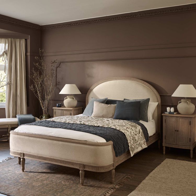 Cozy bedroom with a beige upholstered bed, blue and gray pillows, wooden nightstands, lamps, and a brown accent wall. Large window with curtains.