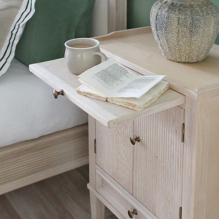 A wooden nightstand with an extended tray holding an open book and a mug, next to a bed with green and white bedding.