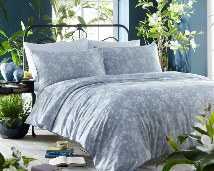 A cozy bedroom with a floral-patterned bedspread, surrounded by lush green plants, books, and soft natural light from a window.
