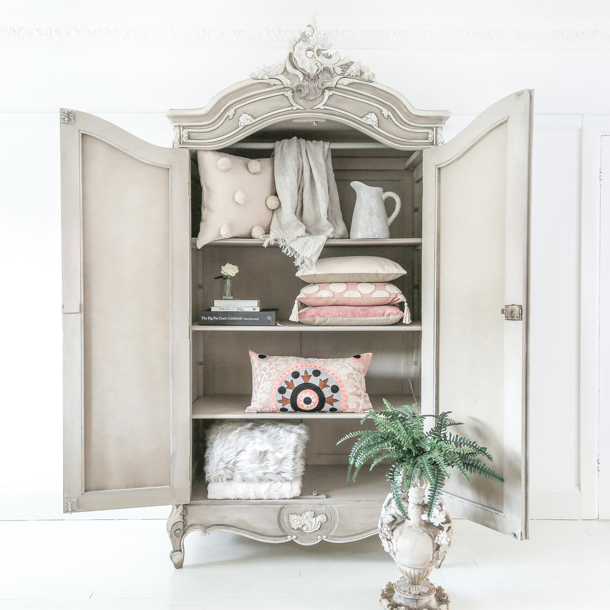 Ornate vintage cabinet with open doors, displaying neatly arranged pillows, a jug, books, and a potted fern below.