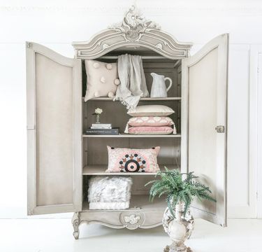 Ornate vintage cabinet with open doors, displaying neatly arranged pillows, a jug, books, and a potted fern below.