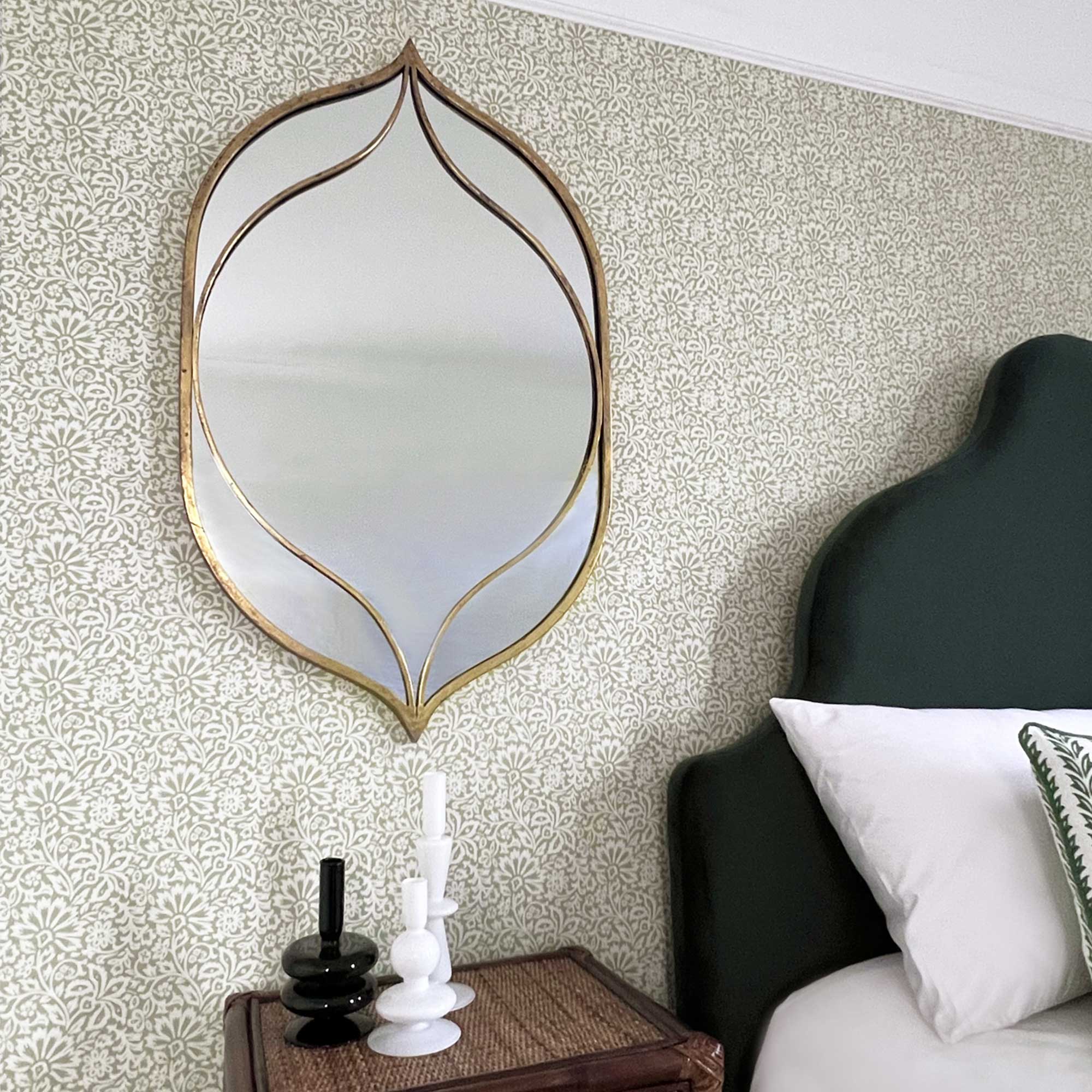 Elegant mirror with gold frame on patterned wallpaper, beside a dark green headboard and a nightstand with candles.