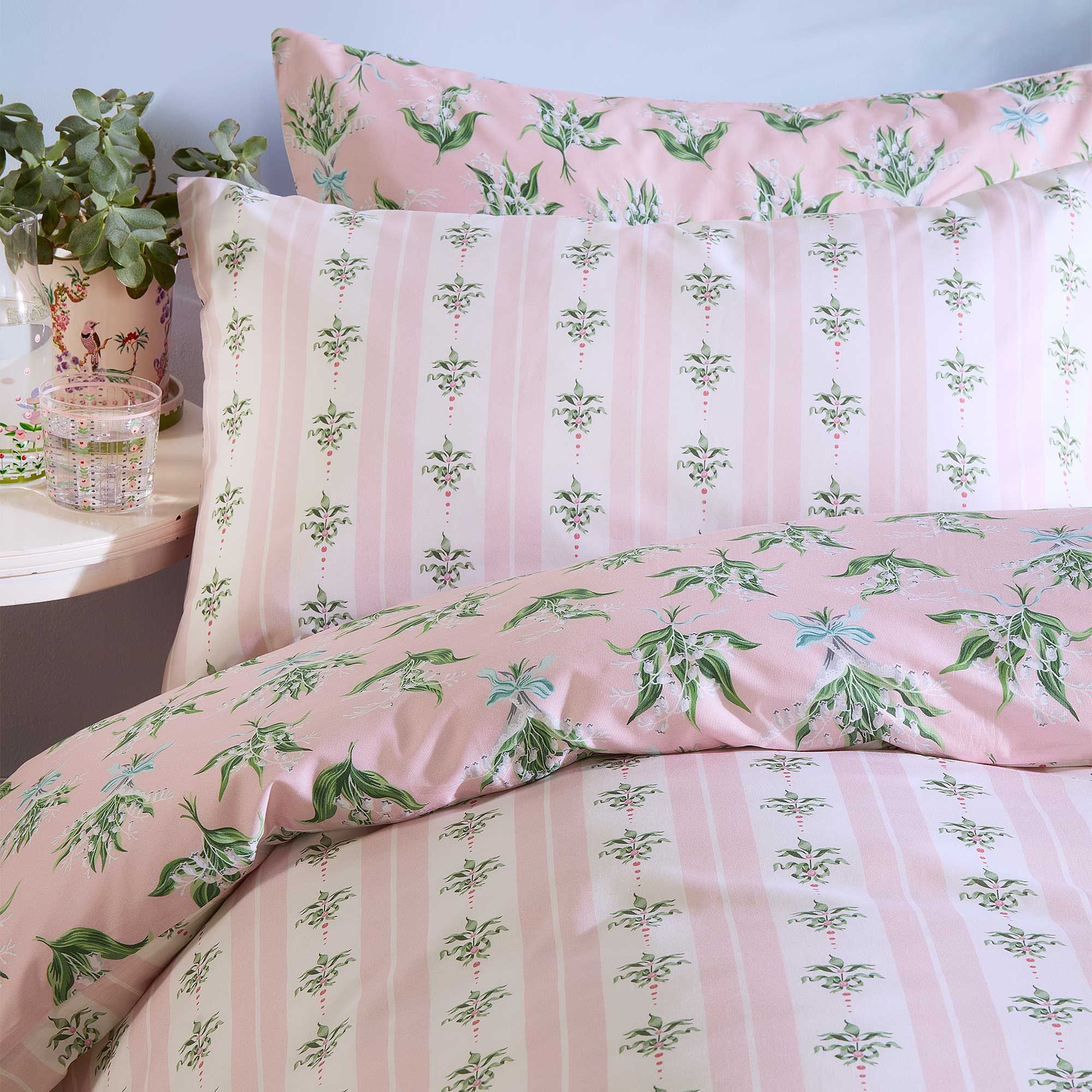 Pink and white striped bedding with green floral patterns, accompanied by a small plant and a patterned glass on a bedside table.