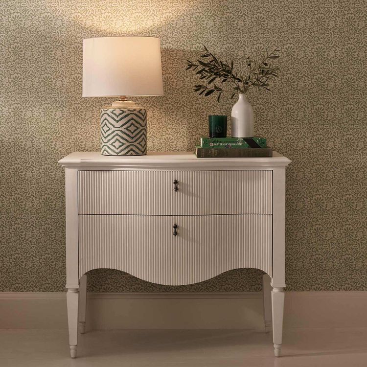 White dresser with two drawers, a patterned lamp, books, and a vase with branches, set against a patterned wallpaper background.