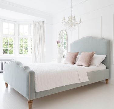 Elegant bedroom with a light blue velvet upholstered bed, white bedding, pink pillows, a chandelier, and a large window with curtains.