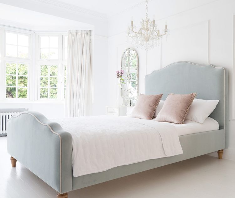 Elegant bedroom with a light blue velvet upholstered bed, white bedding, pink pillows, a chandelier, and a large window with curtains.