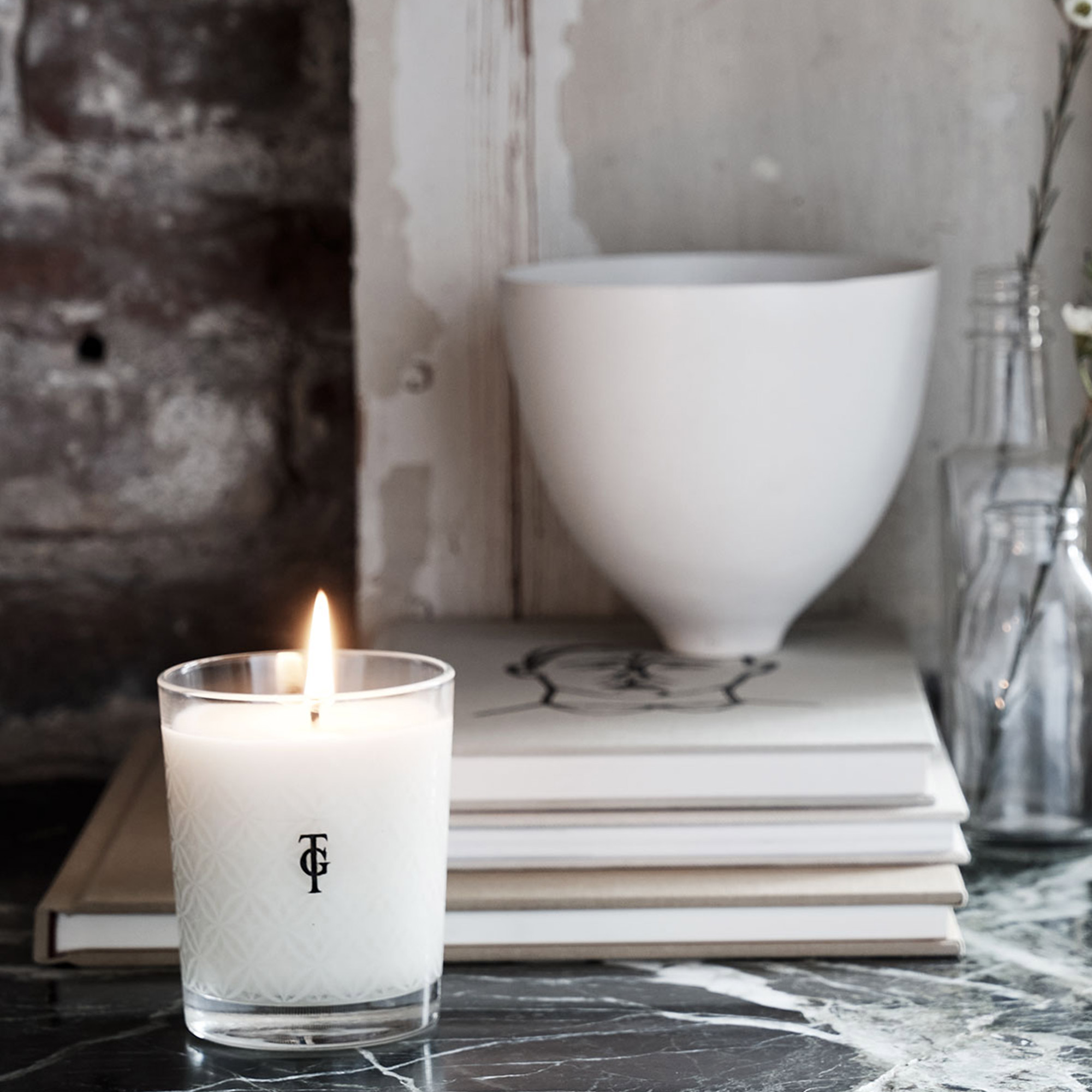 Lit candle in a glass holder on a marble surface, with stacked books, a white bowl, and glass bottles against a textured wall background.
