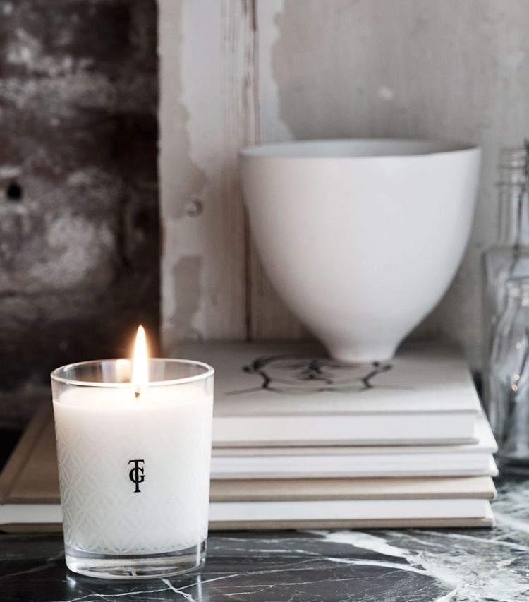 Lit candle in a glass holder on a marble surface, with stacked books, a white bowl, and glass bottles against a textured wall background.