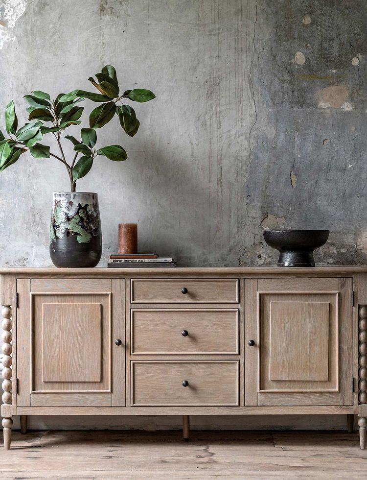 Wooden sideboard with drawers and cabinets, topped with a plant, vase, and bowl, against a textured, weathered wall. Rustic decor.