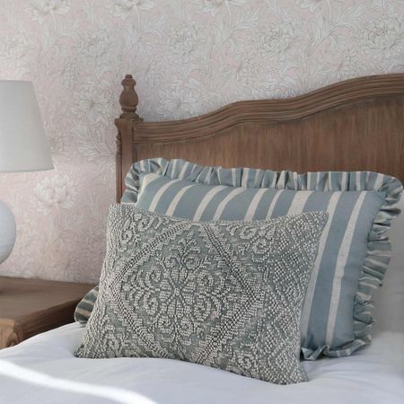 Wooden headboard with floral wallpaper, striped pillow with ruffles, and a textured decorative pillow on a bed. Table lamp on the side.