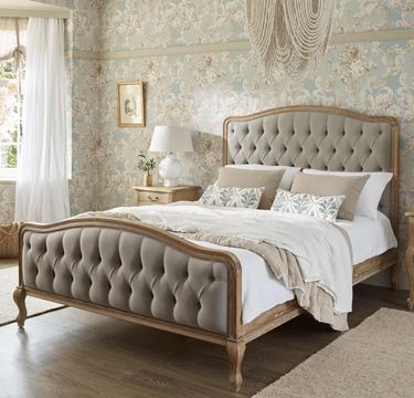 Elegant bedroom with a tufted bed, patterned wallpaper, chandelier, and natural light from a window with sheer curtains.
