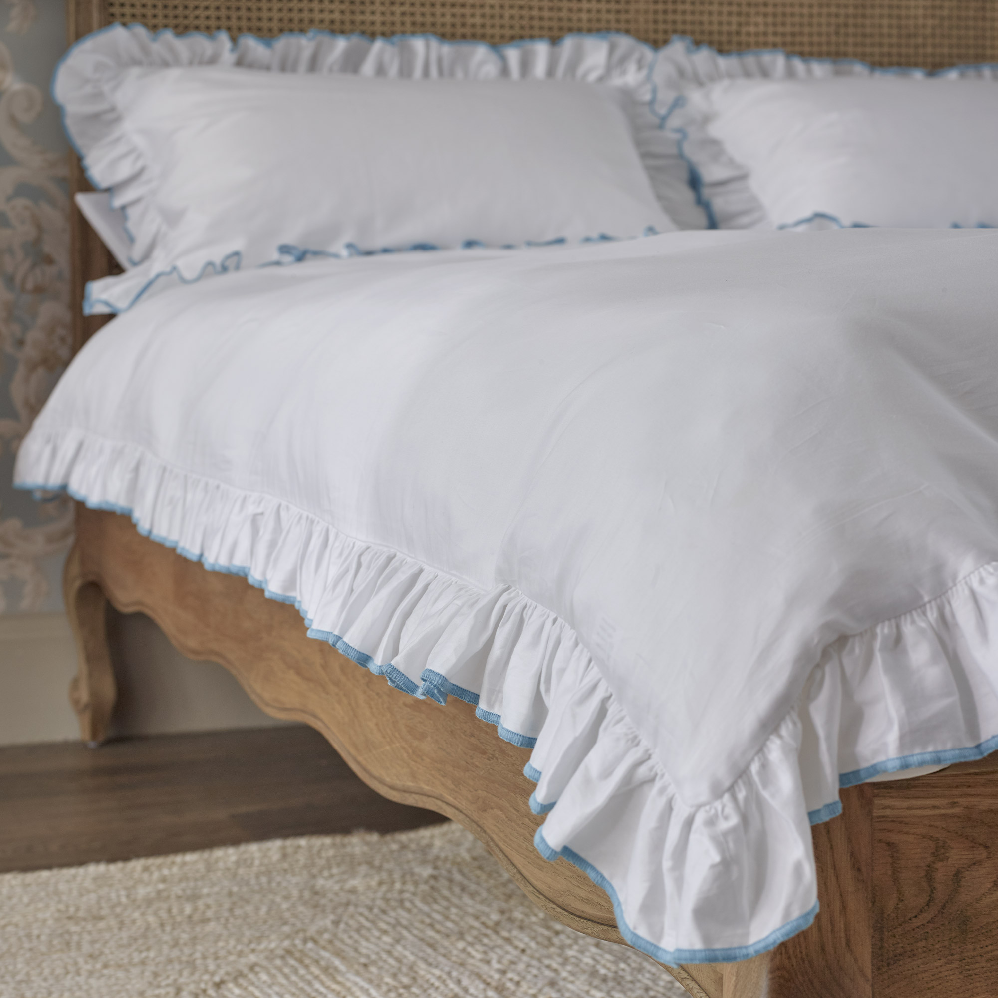 A neatly made bed with white ruffled bedding and blue trim, set on a wooden frame, in a room with floral wallpaper and a light-colored rug.