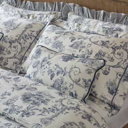Elegant white and blue toile de Jouy bedding featuring intricate floral and pastoral scenes, accented with striped ruffles and piping.