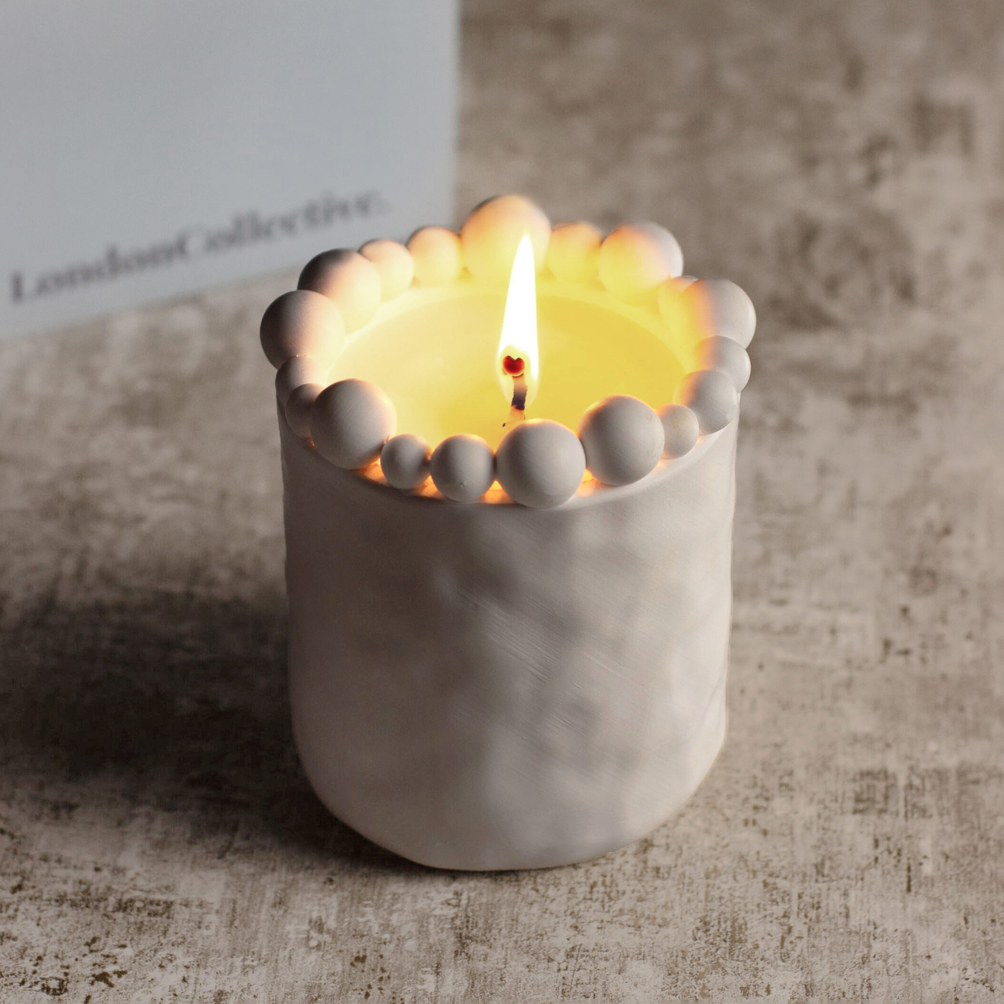 A lit candle in a white holder adorned with round beads on top, placed on a textured surface.