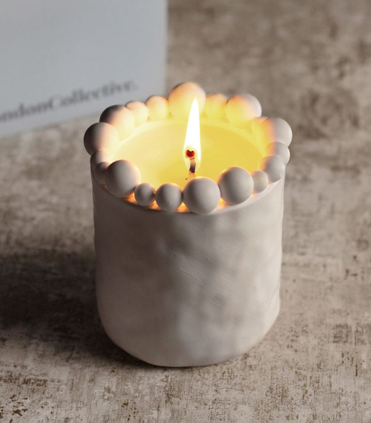 A lit candle in a white holder adorned with round beads on top, placed on a textured surface.