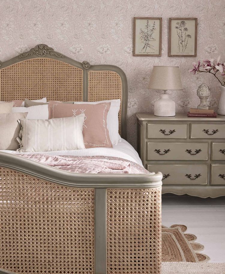 Vintage-style bedroom with a wicker bed, floral wallpaper, and a dresser with a lamp and framed art. Soft pink and beige tones dominate the decor.