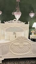 Ornate white bed with intricate carvings, flanked by nightstands and lamps, under elegant chandeliers against a dark green wall.