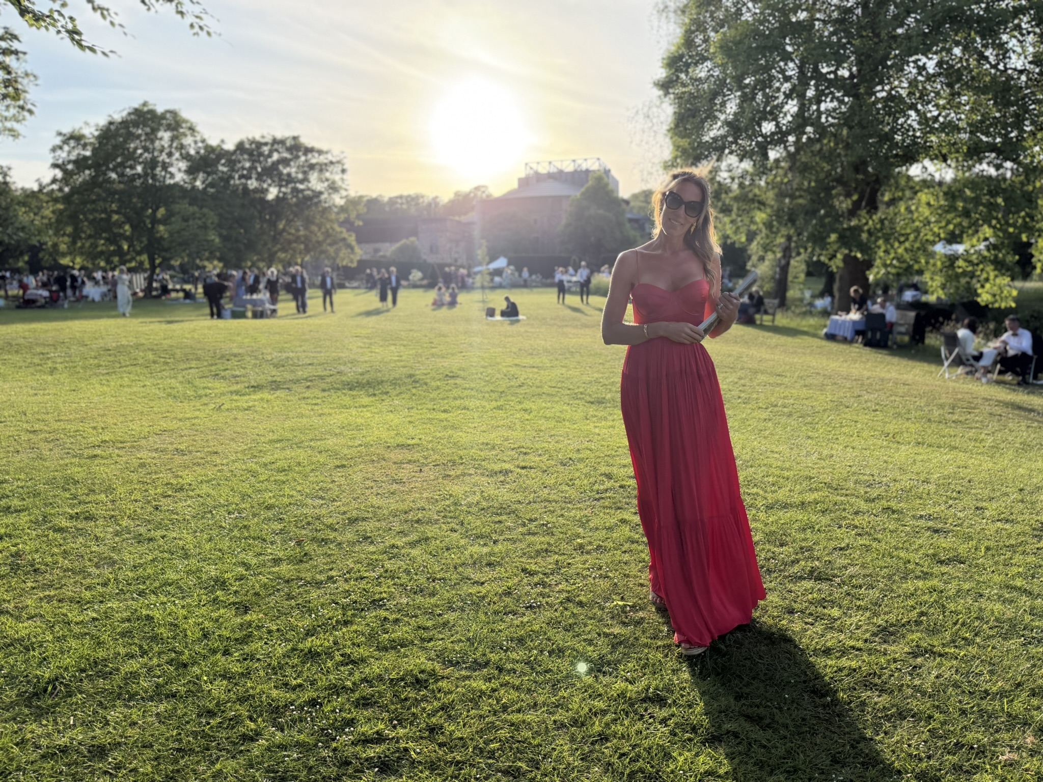 Georgia Metcalfe, Founder and creative director of French Bedroom, in a red dress and sunglasses stands on a sunny lawn outside Glyndebourne Opera House, among trees and picnic setups.