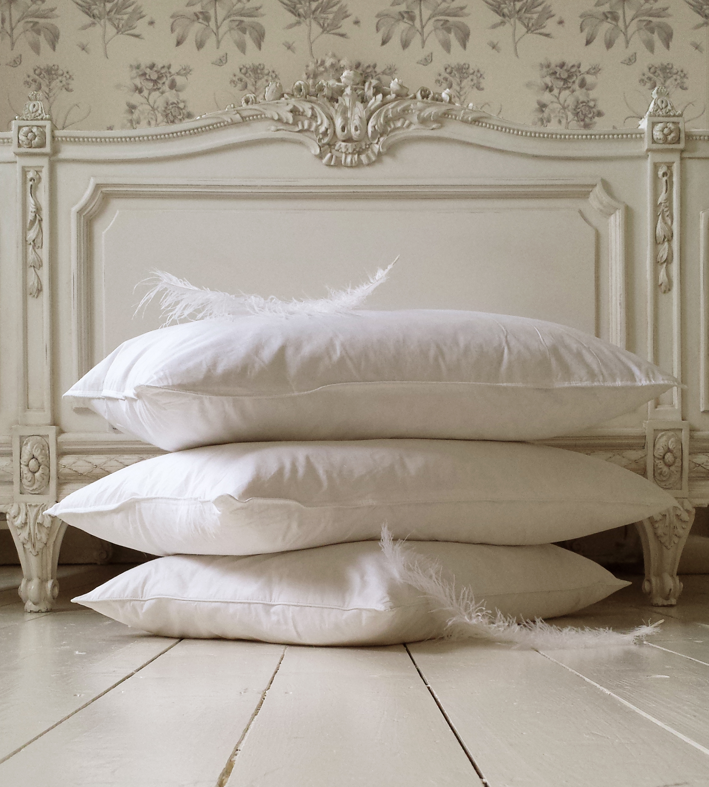 A stack of three white pillows with feathers, placed on a light wooden floor in front of an ornate, vintage white headboard.