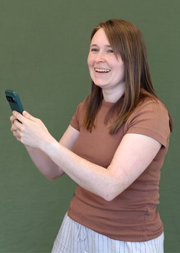 A woman with long brown hair, wearing a brown shirt and striped pants, smiles while holding a smartphone against a green background.