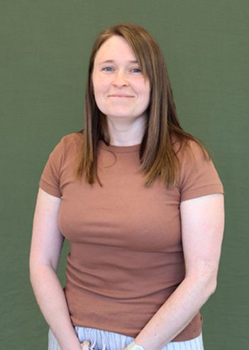 Smiling woman with long brown hair, wearing a brown t-shirt and striped pants, stands against a plain green background.