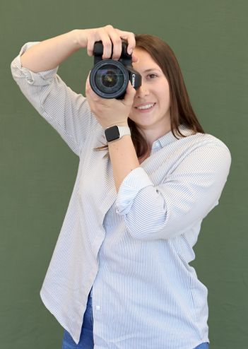 A woman with brown hair in a striped shirt holds a camera to her eye, smiling, against a plain green background.