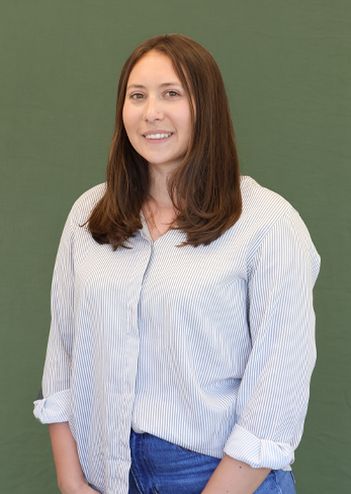 A person with long brown hair, wearing a striped shirt and jeans, stands smiling against a plain green background.