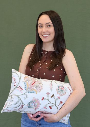 A smiling woman with long dark hair holds an embroidered cushion featuring floral patterns, standing against a green background.