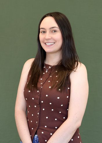 Smiling woman with long dark hair wearing a brown polka dot top stands against a green background.