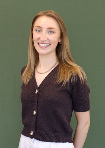 A smiling woman with long blonde hair wearing a dark buttoned shirt and white pants stands in front of a green background.