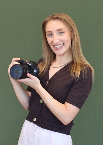Woman smiling, holding a camera, stands against a green background, wearing a brown top and white skirt.