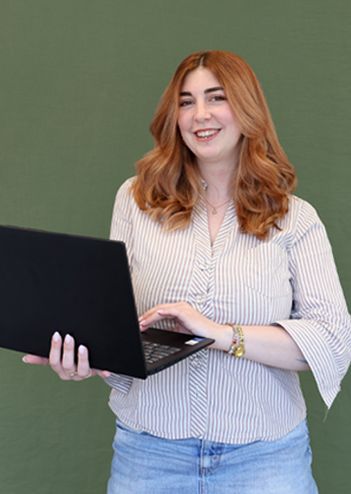 Smiling woman with long hair holding a laptop, standing against a green background.