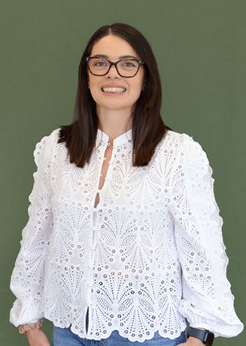 A woman with glasses smiles, wearing a white embroidered blouse and blue jeans, standing against a green background.