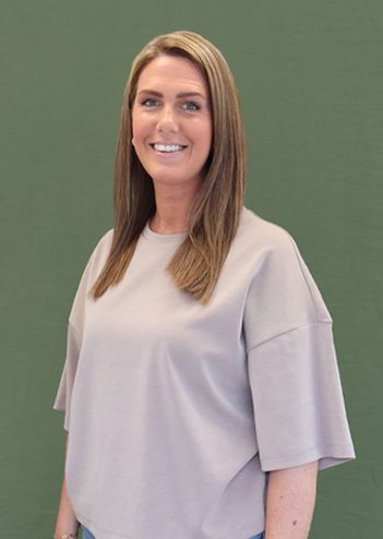 A woman with long brown hair wearing a light grey shirt stands smiling against a green background.