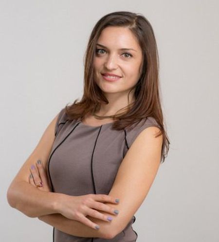 Woman with long brown hair and a gray dress stands with arms crossed, smiling against a plain background.