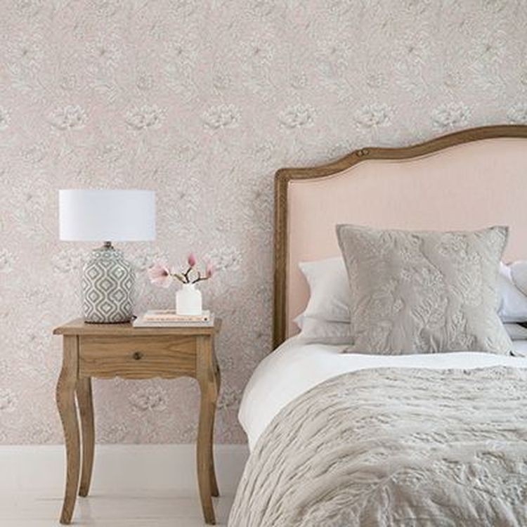 Bedroom scene with a wooden side table, lamp, and vase of flowers. Bed has textured pillows and a quilt, set against floral-patterned wallpaper.