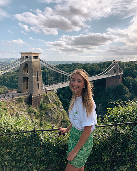 Woman smiling in front of the Clifton Suspension Bridge, surrounded by greenery and a partly cloudy sky.