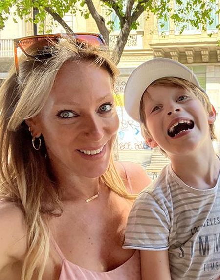 Woman and young boy smiling outdoors, with trees and a building in the background. Boy wears a white cap and striped shirt.