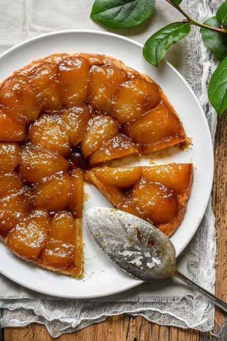 A caramelized apple tarte Tatin on a white plate, with a slice removed. A serving utensil rests nearby on a wooden table.