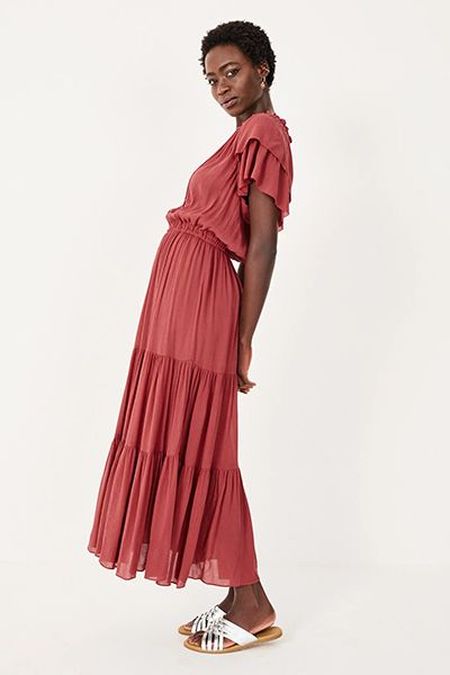 Woman in a flowing, tiered red dress with ruffled sleeves, poses sideways against a white background, wearing white sandals.