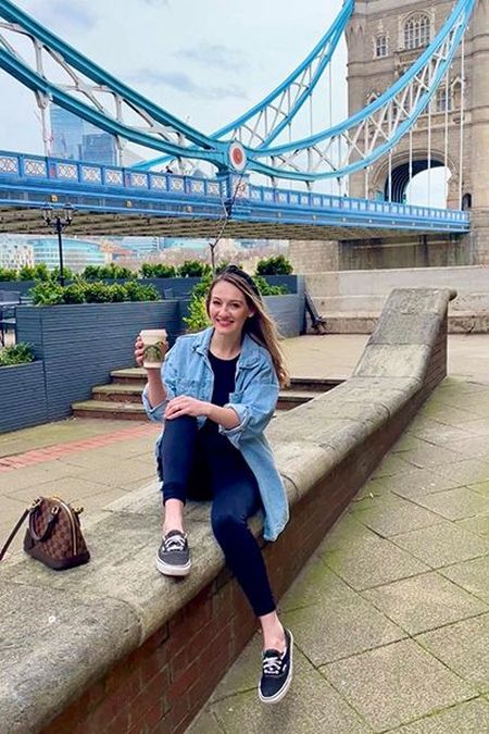 Woman sitting on a stone ledge holding a coffee cup, smiling. Tower Bridge in the background. Casual attire, sunny day.