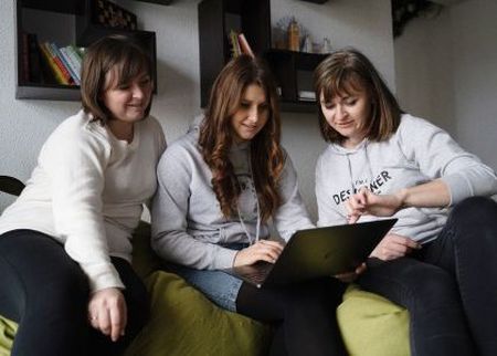 Three women sitting on a couch, engaged with a laptop. Two are in hoodies, and one in a sweater. Shelves with books are in the background.