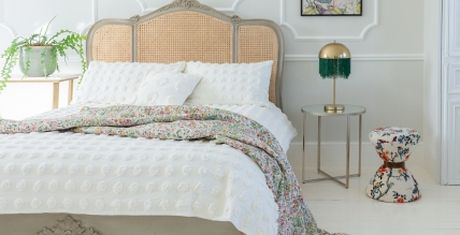 Elegant bedroom with a vintage-style bed, textured white bedding, floral throw, gold lamp on a glass table, and a floral-patterned stool.