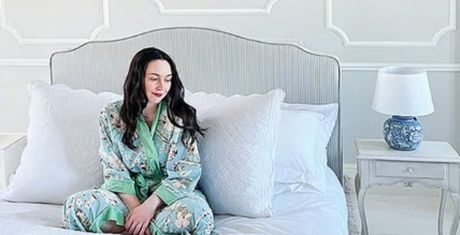 A person in floral pajamas sits on a white bed with large pillows, next to a nightstand with a blue and white lamp, in a bright room.