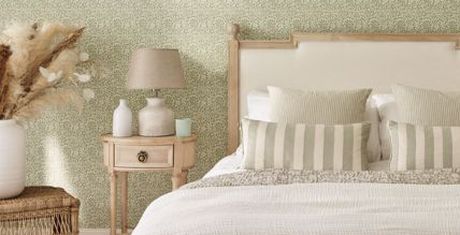 Cozy bedroom with a patterned green wallpaper, a wooden bed with striped pillows, a side table with vases, and a lamp.