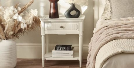 A cozy bedroom scene with a white nightstand, decorative vases, pampas grass, and a neatly made bed with a soft beige blanket.