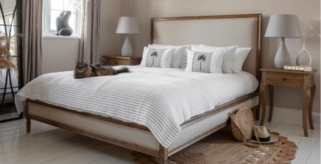 Cozy bedroom with a large bed, palm-themed pillows, a cat lounging, and rustic wooden furniture. A straw hat and bag are on the floor.