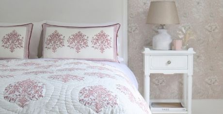 Cozy bedroom with a bed featuring floral-patterned bedding, matching pillows, and a white nightstand with a lamp and decor against floral wallpaper.