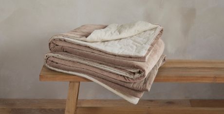 Folded beige and cream quilts stacked on a wooden bench against a neutral wall.