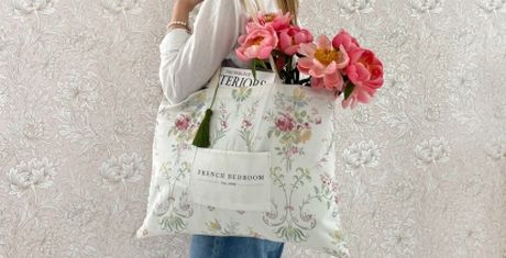 Woman holding a floral tote bag with pink flowers and a magazine inside, standing against a patterned wallpaper.